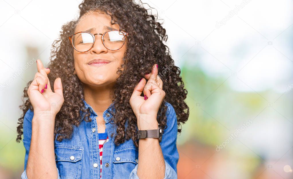 Young beautiful woman with curly hair wearing glasses smiling crossing fingers with hope and eyes closed. Luck and superstitious concept.