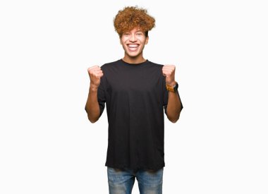 Afro saçlı siyah kutluyor t-shirt giyen genç yakışıklı adam şaşırttı ve başarı için kaldırdı kollarını açarak hayret ve gözleri açık. Kazanan kavramı.