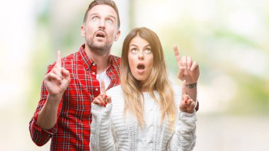 Aşık izole arka plan üzerinde kış kazak giyen genç çift hayrete ve yukarıya bakıyor ve parmakları ile işaret ve silah kaldırdı şaşırttı.