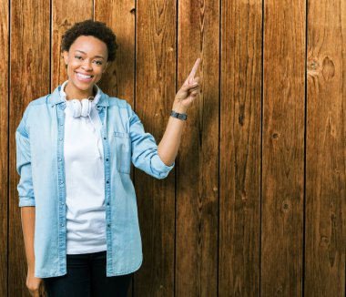 Kameraya bakarak yan el ve parmak ile işaret yüzünde kocaman bir gülümseme ile izole arka plan üzerinde kulaklık giyen genç Afro-Amerikan kadın.