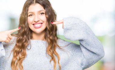 Kendinden emin gösterilen ve işaret parmakları diş ve ağız ile gülümseyen kış kazak giyen genç güzel kadın. Sağlık kavramı.