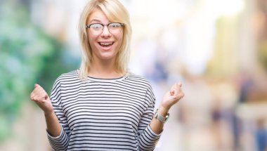 Genç güzel sarışın kadın izole arka plan üzerinde kutluyor gözlük takan şaşırttı ve başarı için kaldırdı kollarını açarak hayret ve gözleri açık. Kazanan kavramı.