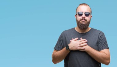 Genç beyaz hipster adam kapalı gözler ile göğüs ellerinde ve yüzünde minnettar jest ile gülümseyen izole arka plan üzerinde güneş gözlüğü takıyor. Sağlık kavramı.