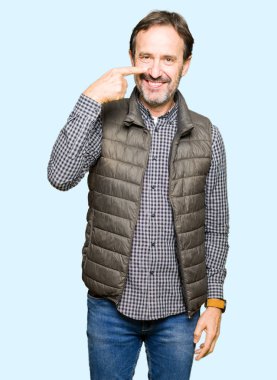 Orta yaş yakışıklı adam giyen kış yüz ve burun, neşeli gülümseyerek el parmak ile işaret yelek