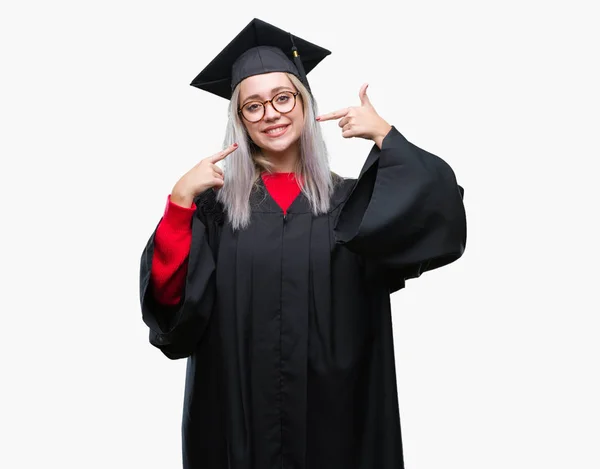 年轻的金发碧眼的女人穿着研究生制服在孤立的背景微笑自信显示和指向用手指的牙齿和嘴 健康理念 — 图库照片