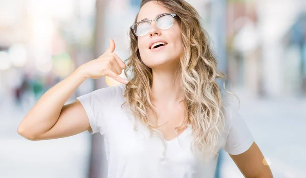 Beautiful young blonde woman wearing glasses over isolated background smiling doing phone gesture with hand and fingers like talking on the telephone. Communicating concepts.