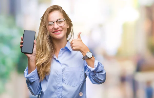 年轻美丽的金发碧眼的女商人显示屏幕上的智能手机在孤立的背景高兴与大微笑做确定的标志 用手指竖起大拇指 优秀的标志 — 图库照片