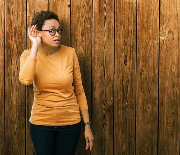 Young beautiful african american woman wearing glasses over isolated background smiling with hand over ear listening an hearing to rumor or gossip. Deafness concept.