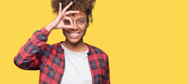 Beautiful young african american woman wearing glasses over isolated background doing ok gesture with hand smiling, eye looking through fingers with happy face.