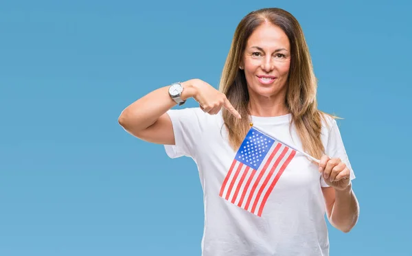 Middle age hispanic woman holding flag of United States of America over isolated background with surprise face pointing finger to himself
