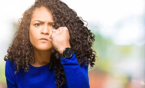 Young beautiful woman with curly hair wearing winter sweater angry and mad raising fist frustrated and furious while shouting with anger. Rage and aggressive concept.