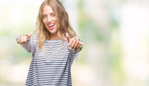 Beautiful young blonde woman wearing stripes sweater over isolated background approving doing positive gesture with hand, thumbs up smiling and happy for success. Looking at the camera, winner gesture.