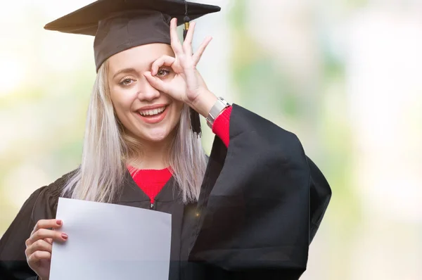 指の間から見て目に手で サインをして笑っている幸せそうな顔で孤立の背景の上の学位を保持する大学院の制服を着て若いブロンドの女性 — ストック写真