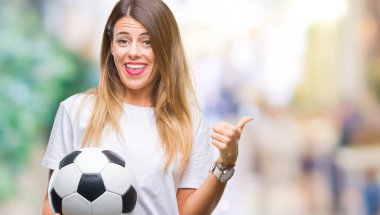 İşaret ve başparmak kadar mutlu yüz gülümseyen tarafı ile gösteren izole arka plan üzerinde futbol topu tutan genç güzel kadın