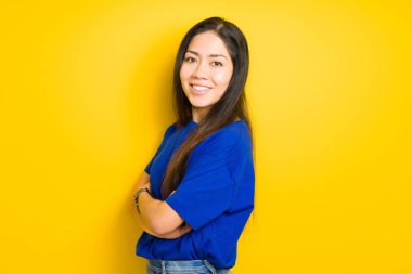 Çapraz kolları ile ayakta ve sarı izole arka plan üzerinde gülümseyen güzel genç esmer kadın