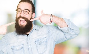 Uzun saç ve sakal emin gösterilen ve işaret parmakları diş ve ağız ile gülümseyen gözlüklü genç hipster adam. Sağlık kavramı.