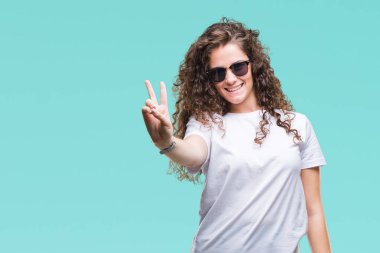 Güzel esmer kıvırcık saçlı Genç kız mutlu yüz zafer işareti yaparken kameraya göz kırpıyor ile gülümseyen izole arka plan üzerinde güneş gözlüğü takıyor. İki numaralı.