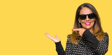 Güzel orta yaş kadın hayrete ve el ile sunulması ve parmak ile işaret ederken kameraya gülümseyen güneş gözlüğü takıyor.