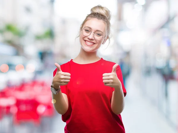 Young blonde woman wearing glasses over isolated background approving doing positive gesture with hand, thumbs up smiling and happy for success. Looking at the camera, winner gesture.