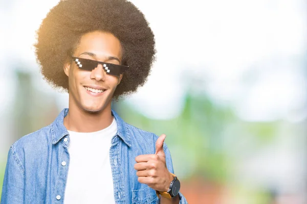 Young african american man with afro hair wearing thug life glasses doing happy thumbs up gesture with hand. Approving expression looking at the camera with showing success.