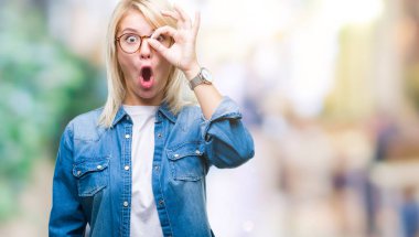 Sürpriz yüz, parmaklarının arasından seyir göz gözlük ok jest yapıyor izole arka plan üzerinde giyen genç güzel sarışın kadın şok. İmansız ifade.