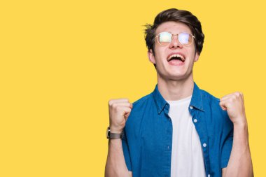 Yakışıklı delikanlı izole arka plan üzerinde kutluyor gözlük takan şaşırttı ve başarı için kaldırdı kollarını açarak hayret ve gözleri açık. Kazanan kavramı.