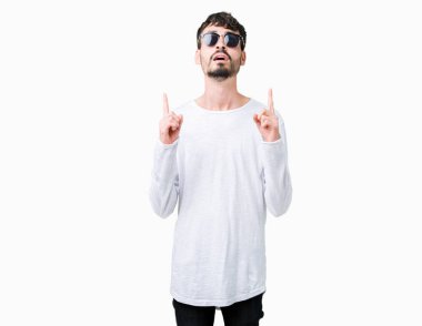 Yakışıklı delikanlı hayrete ve yukarıya bakıyor ve parmakları ile işaret ve silah kaldırdı sürpriz izole arka plan üzerinde güneş gözlüğü takıyor.