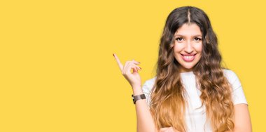 Kameraya bakarak yan el ve parmak ile işaret, yüzünde kocaman bir gülümseme ile rahat beyaz t-shirt giyen genç güzel kadın.