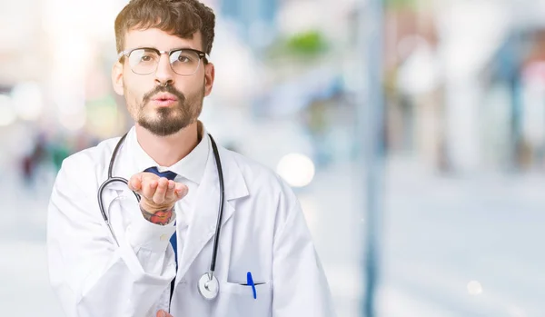 Young doctor man wearing hospital coat over isolated background looking at the camera blowing a kiss with hand on air being lovely and sexy. Love expression.