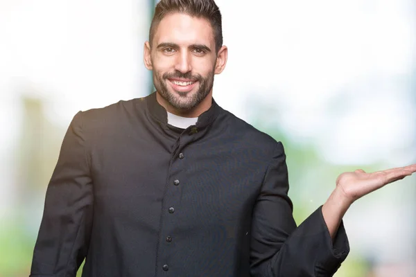 Young Christian priest over isolated background smiling cheerful presenting and pointing with palm of hand looking at the camera.
