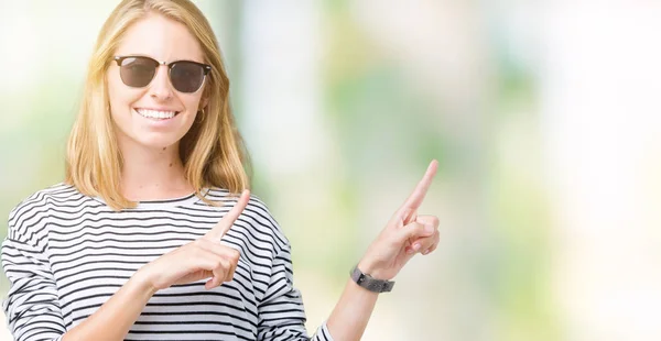 Beautiful young woman wearing sunglasses over isolated background smiling and looking at the camera pointing with two hands and fingers to the side.