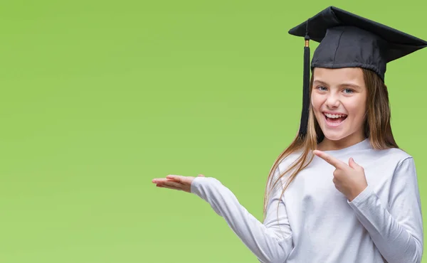 Young beautiful girl wearing graduate cap over isolated background amazed and smiling to the camera while presenting with hand and pointing with finger.