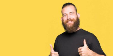 El ile olumlu hareketi yaparken rahat t-shirt başarı işareti giyen genç sarışın adam başparmak gülümseyen ve mutlu. Neşeli bir ifadeyle, kazanan jest kameraya bakıyor.