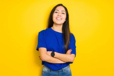 Kameraya bakarak çapraz silah ile gülümseyen izole sarı arka plan mutlu yüzüne mavi t-shirt giyen güzel esmer kadın. Olumlu bir kişi.