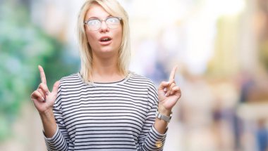 Genç güzel sarışın kadın hayrete ve yukarıya bakıyor ve parmakları ile işaret ve silah kaldırdı sürpriz izole arka plan üzerinde gözlük.