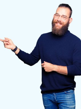 Kameraya bakarak yan el ve parmak ile işaret yüzünde, gözlük ve kocaman bir gülümseme ile balıkçı yaka kazak giyen genç sarışın adam.