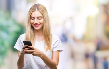 Ayakta ve diş gösterilen kendine güvenen bir gülümsemeyle gülümseyerek mutlu bir yüz ile izole arka plan üzerinde akıllı telefon kullanan genç güzel sarışın kadın