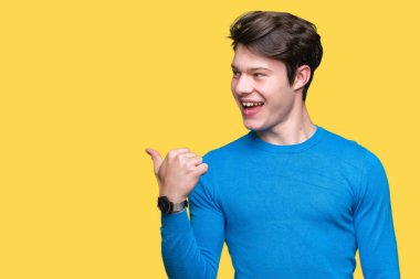 Yakışıklı delikanlı arıyor ve başparmak yanında yukarıyı mutlu yüz ile gülümseyen izole arka plan üzerinde mavi kazak giyiyor.