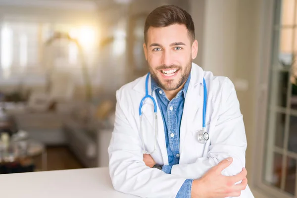 Young handsome doctor man at the clinic happy face smiling with crossed arms looking at the camera. Positive person.