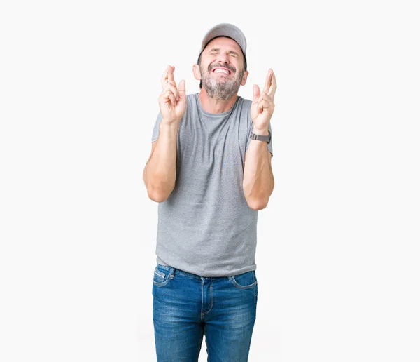 ハンサムな中年白髪シニア男性希望と目を閉じて交差指の笑みを浮かべて隔離された背景にスポーツ キャップを着用します 運と迷信的な概念 — ストック写真