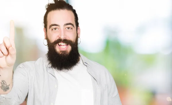 Young man with long hair, beard and earrings showing and pointing up with finger number one while smiling confident and happy.