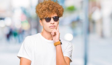 Yakışıklı genç afro saçlı çene düşünme hakkında soru, dalgın ifade üzerine el ile güneş gözlüğü takıyor. Düşünceli yüzüyle gülümsüyor. Şüphe kavramı.