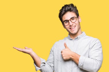 İzole arka plan gösteren palm el üzerinde gözlük takan ve yukarıya, mutlu ve neşeli gülümseyen baş parmakları Tamam jest yapıyor yakışıklı delikanlı