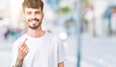 Beckoning gel burada mutlu ve gülümseyerek el davet ile jest izole arka plan üzerinde beyaz t-shirt giyen genç yakışıklı adam