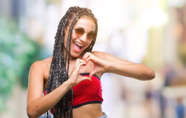 Genç örgülü saç Afrikalı-Amerikalı Doğum işareti kalp simgesi ve elleriyle şekil gösterilen aşık gülümseyen izole arka plan üzerinde güneş gözlüğü takıyor. Romantik kavramı.