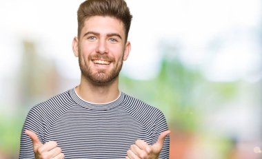 Yakışıklı delikanlı çizgili süveter başarı işareti el ile olumlu hareketi yaparken giyiyor başparmak gülümseyen ve mutlu. Neşeli bir ifadeyle, kazanan jest kameraya bakıyor.