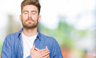 Genç yakışıklı sarışın adam rahat denim ceket ile gülümseyen giyen kapalı gözler ve yüzünde minnettar jest ile göğüs üzerinde eller. Sağlık kavramı.