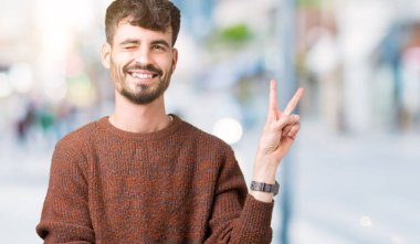 Mutlu yüz zafer işareti yaparken kameraya göz kırpıyor ile gülümseyen izole arka plan üzerinde kış kazak giyen genç yakışıklı adam. İki numaralı.