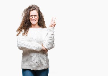 Güzel esmer kıvırcık saçlı Genç kız mutlu yüz zafer işareti yaparken kameraya göz kırpıyor ile gülümseyen izole arka plan üzerinde kış kazak giyiyor. İki numaralı.