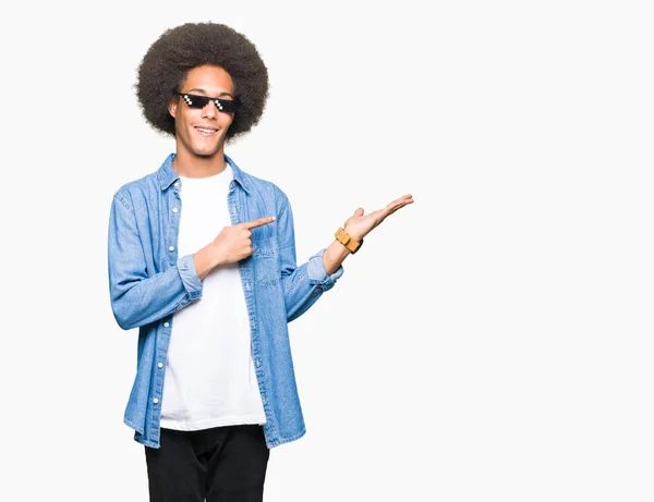 Young african american man with afro hair wearing thug life glasses amazed and smiling to the camera while presenting with hand and pointing with finger.